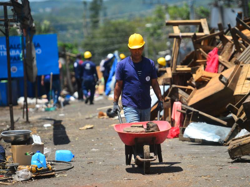 Trabajador con casco amarillo empuja una carretilla cargada con pedazos de concreto frente a un gran montón de tarimas de madera.