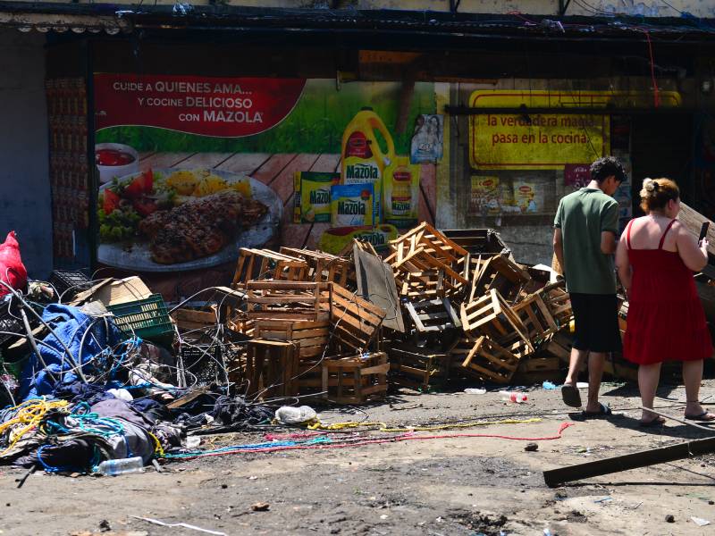 Un hombre y una mujer caminan sobre la calle frente a una pila de cajas de madera y residuos de puestos desalojados.