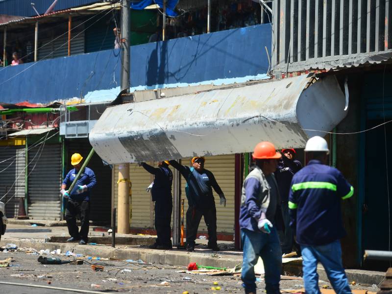 Grupo de operarios levantando y desmontando un techo o marquesina de metal frente a los locales comerciales en la acera.