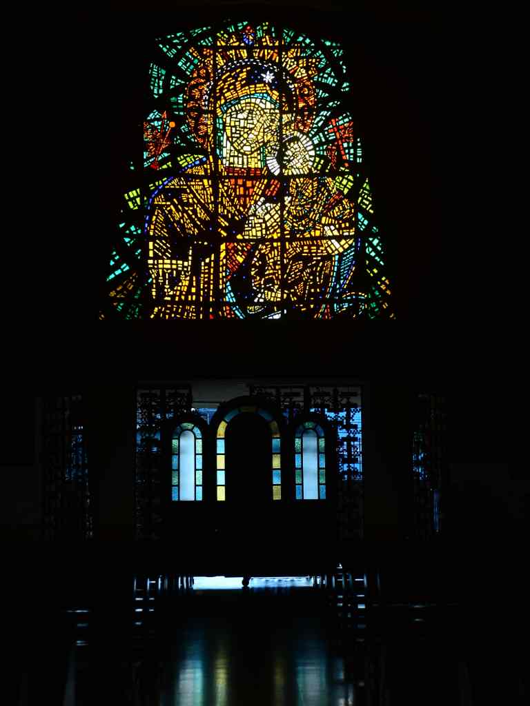 Vitral luminoso en forma de arco que representa a la Virgen con el Niño Jesús al fondo de la nave central a oscuras.