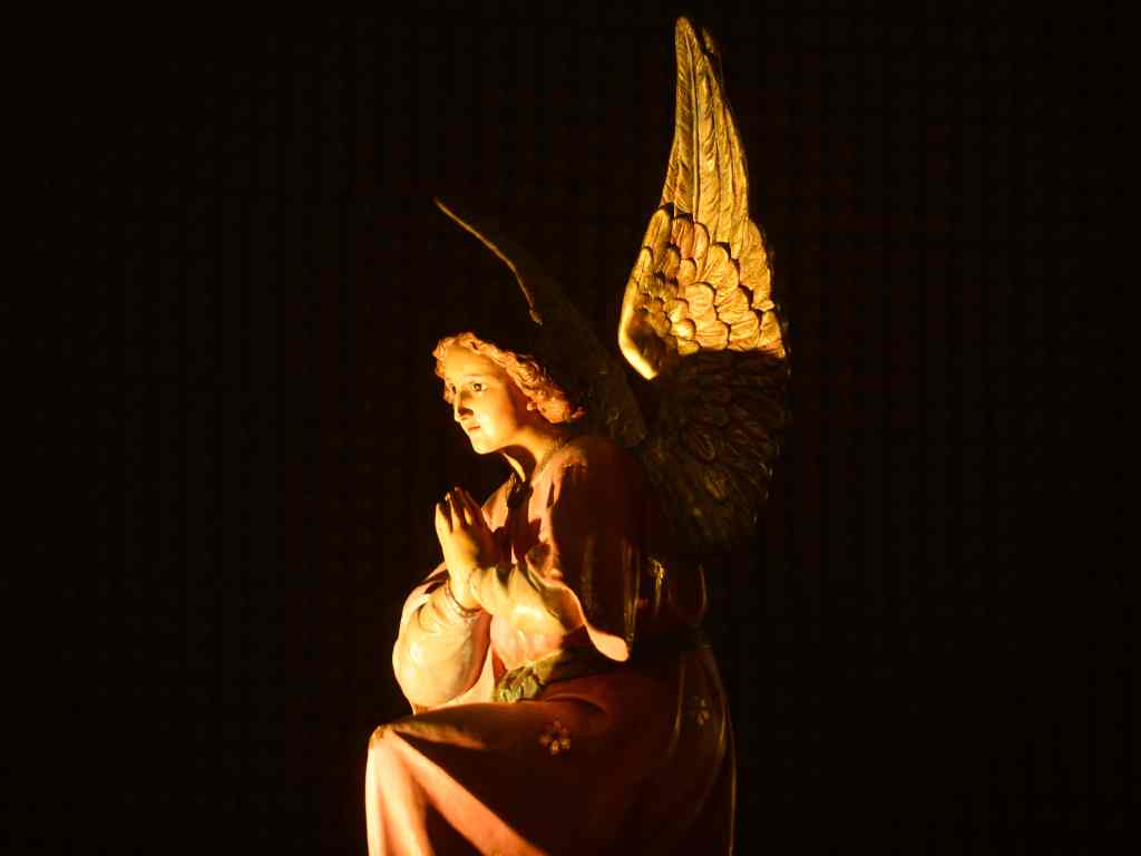 Escultura de un ángel arrodillado con las manos juntas en gesto de oración y vestimenta túnica bajo luz directa.