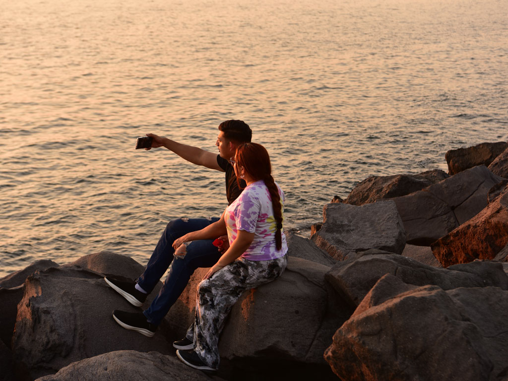 Pareja sentada sobre las rocas de la costa tomándose una fotografía con su teléfono móvil al atardecer.