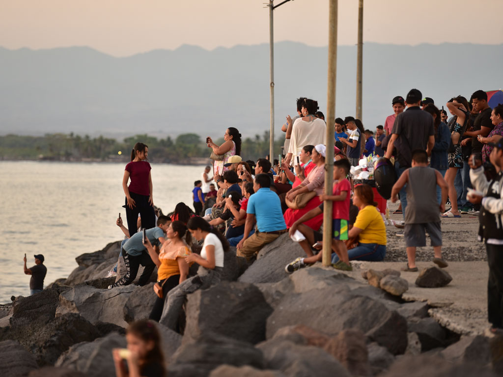 Una fotografía de ángulo medio de un gran grupo de personas reunidas en un malecón rocoso al atardecer, mirando hacia el océano y las montañas lejanas.