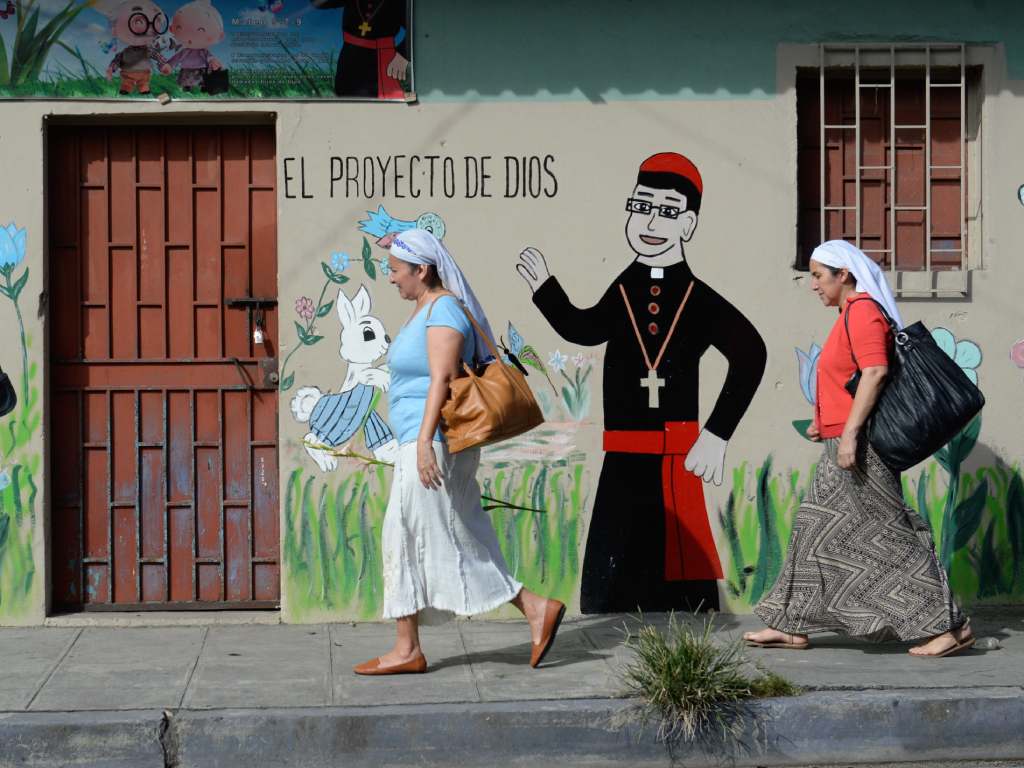 Dos mujeres caminan frente a un colorido mural de Romero con estilo de caricatura.
