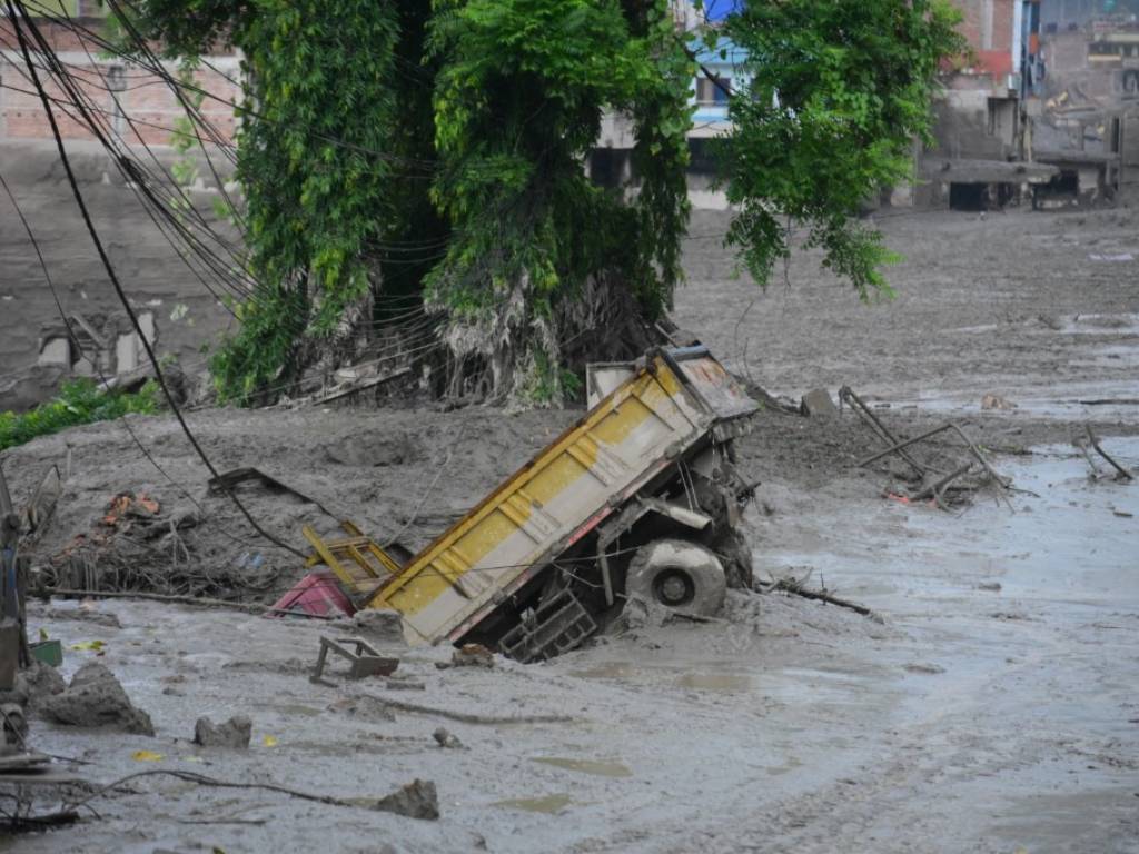 Vehículo pesado atrapado e inclinado en lodo gris. Cabina sumergida, ruedas visibles, edificios dañados y cables colgando