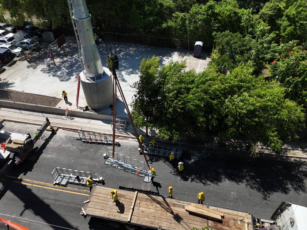 Vista aérea de trabajadores supervisando el izaje de piezas metálicas con grúa junto a la base de la torre.