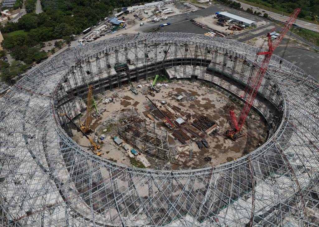 Vista aérea en picado de la obra del Estadio Nacional de El Salvador. Se observa una inmensa armazón metálica de techo circular envolviendo las graderías de concreto y varias grúas de construcción operando en el terreno central de tierra.