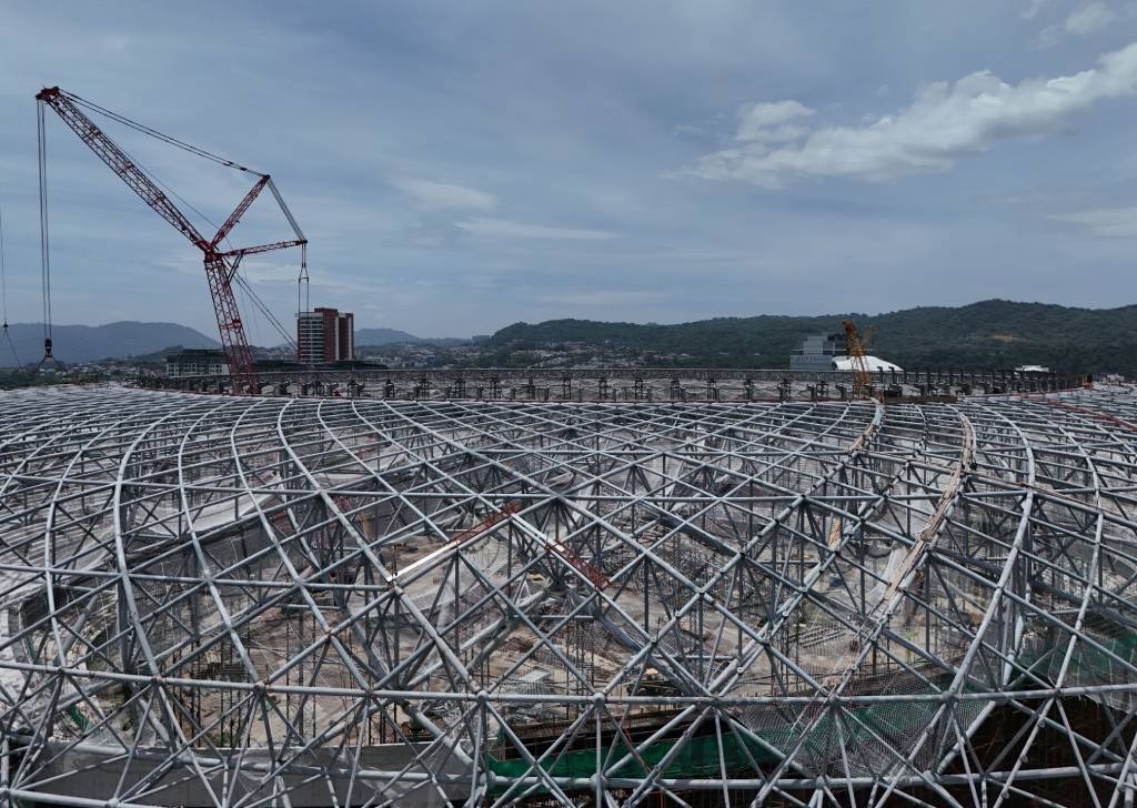 Toma aérea del techo en construcción del estadio, dominado por una tupida red de vigas metálicas plateadas. A la izquierda destaca una enorme grúa roja alzándose sobre la estructura, mientras al fondo se aprecian edificios urbanos y montañas bajo un cielo despejado.