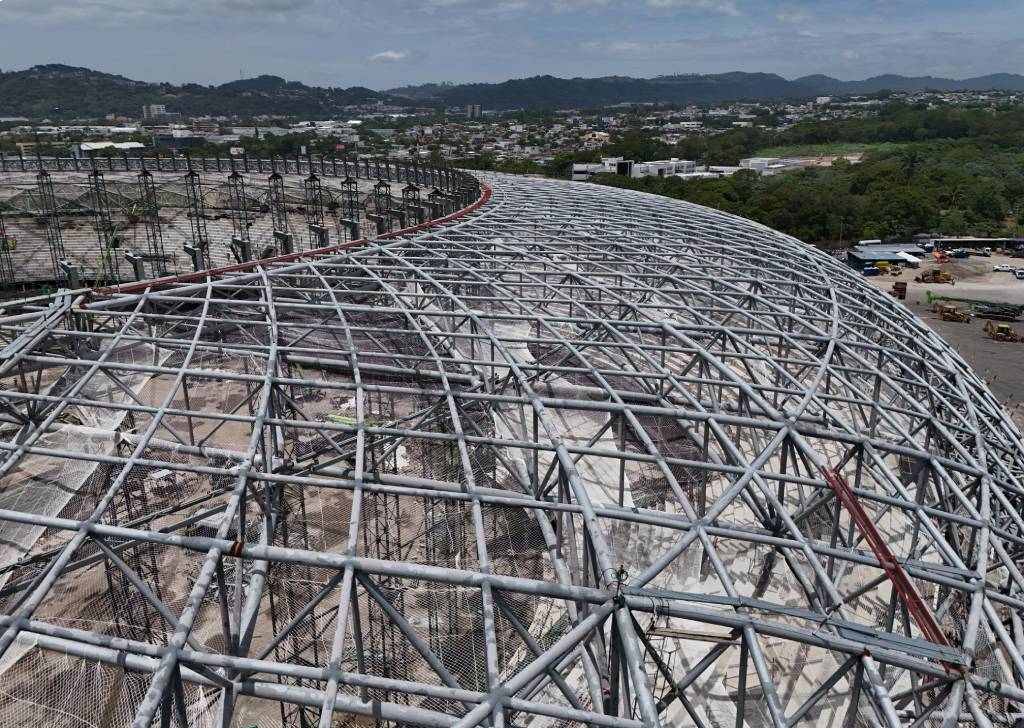 Primer plano aéreo de la intrincada estructura de red de acero plateado en construcción para el techo del estadio. Se aprecian los múltiples puntos de conexión y la curvatura de la forma. El fondo muestra la ciudad y las colinas bajo un cielo parcialmente nublado.