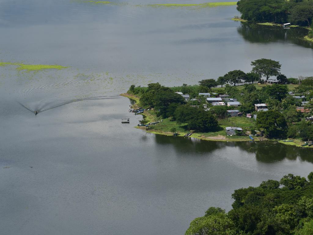 Casas rústicas con techos de lámina se agrupan en la orilla verde, con botes amarrados cerca.