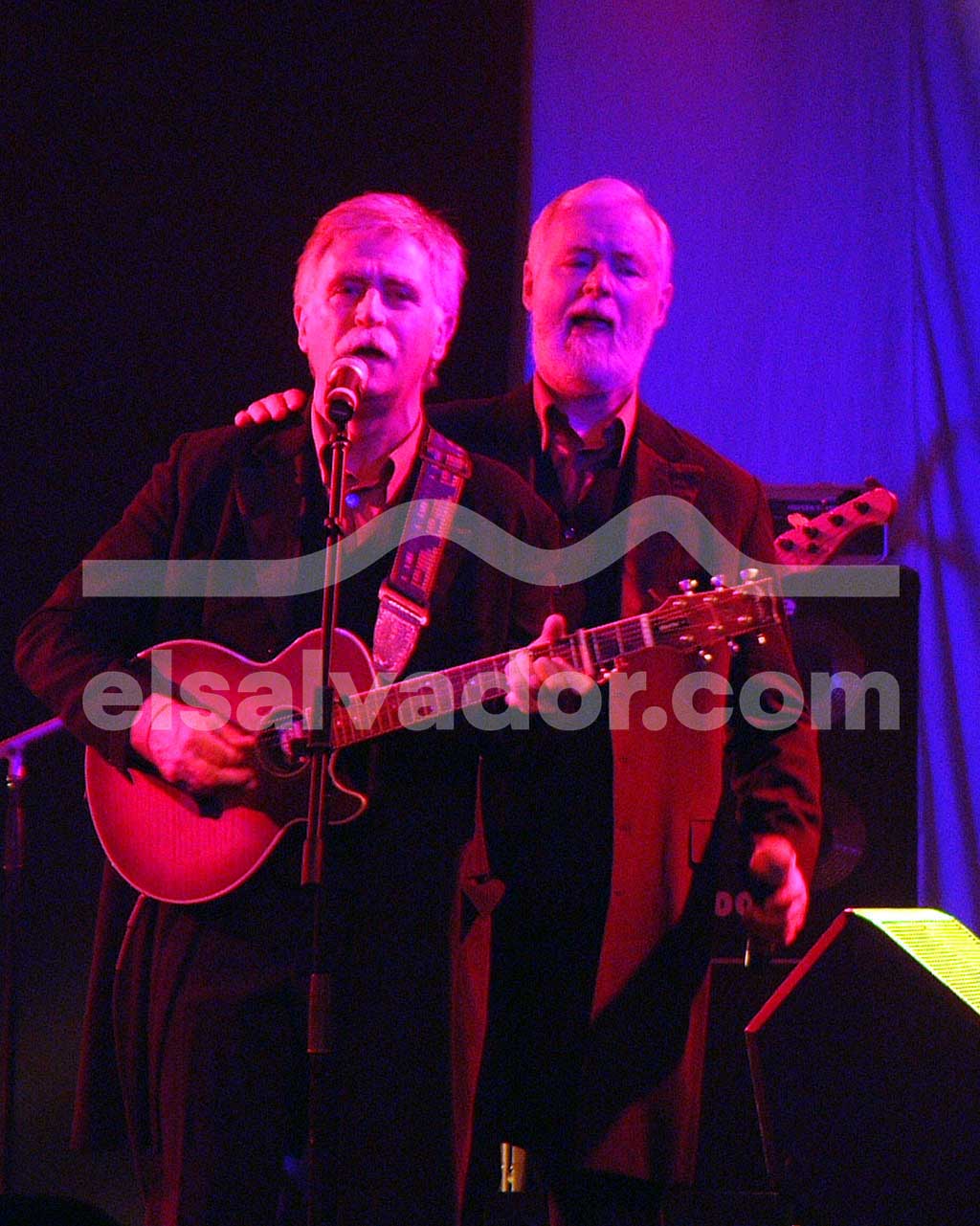 Dos cantantes masculinos de El Consorcio en el escenario; uno toca la guitarra acústica mientras el otro canta a su lado bajo luces moradas