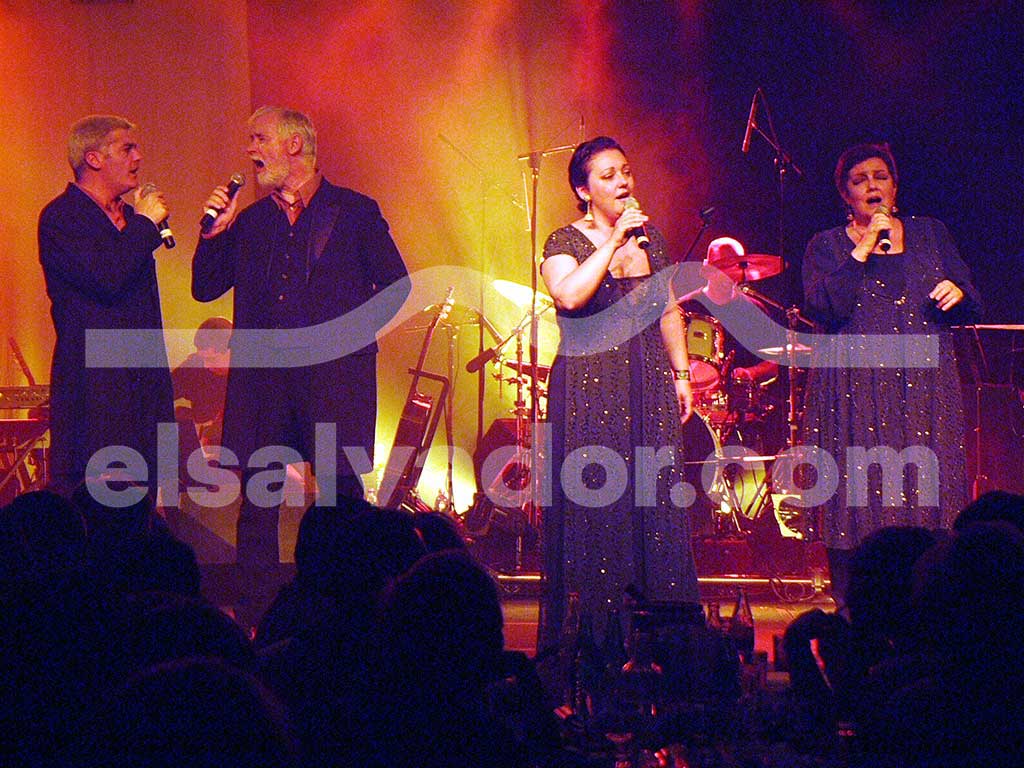 Cuatro integrantes de El Consorcio cantando apasionadamente en micrófonos bajo reflectores cálidos en el escenario.