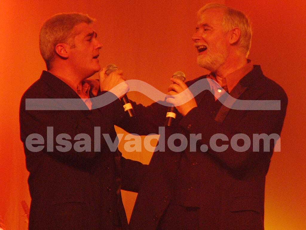 Plano medio de dos cantantes masculinos compartiendo un dueto en el escenario bajo una cálida iluminación roja.
