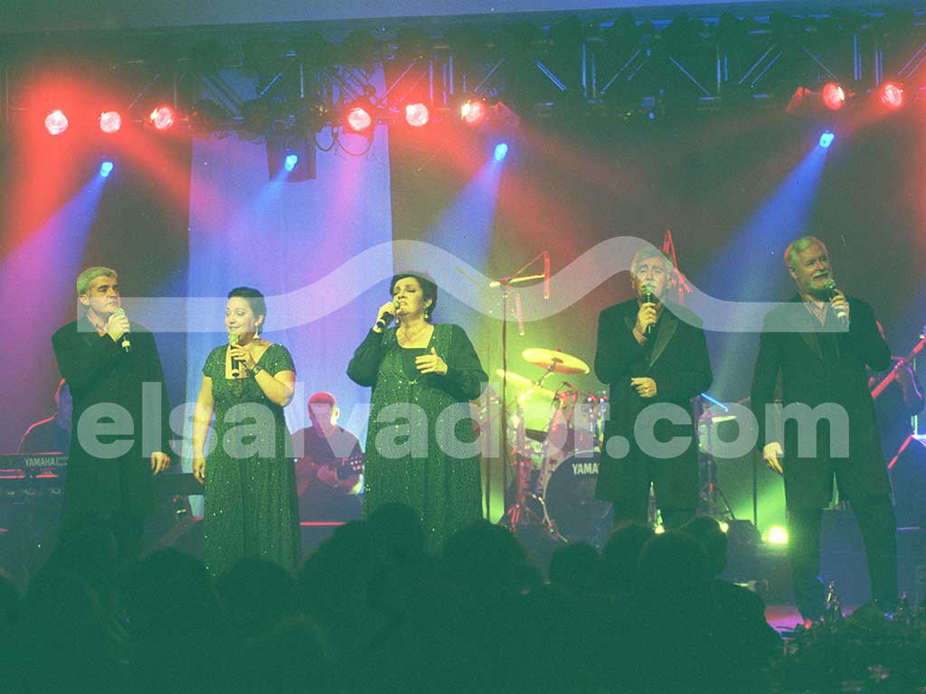 Foto de plano entero de los cinco cantantes de El Consorcio alineados en el escenario cantando juntos entre luces azules y rojas.