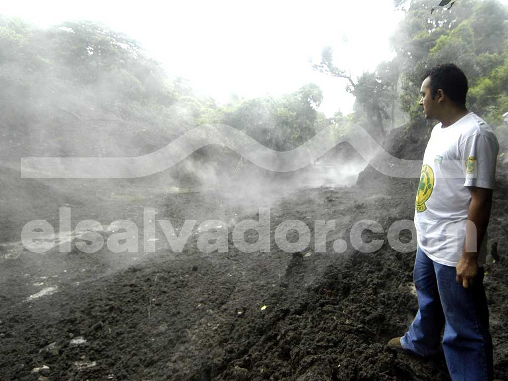 Un hombre con camiseta blanca y la insignia de Comandos de Salvamento contempla un terreno de fango oscuro del que emanan densas columnas de vapor.