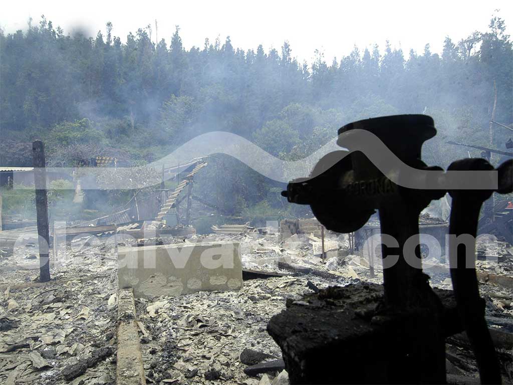 Primer plano en silueta de un molino de maíz de hierro fijado sobre una mesa en ruinas, con el fondo de una casa totalmente destruida por el fuego y la ceniza.