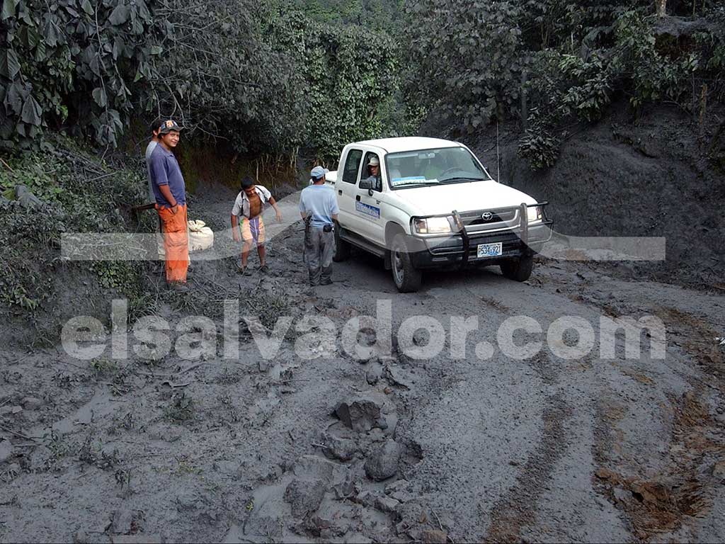 Un pick-up blanco con la marca "COOPERATIVA LOS NARANJOS" en la puerta está atascado en una carretera cubierta de lodo gris espeso y ceniza. Varias personas están de pie cerca del vehículo en el paisaje desolado.