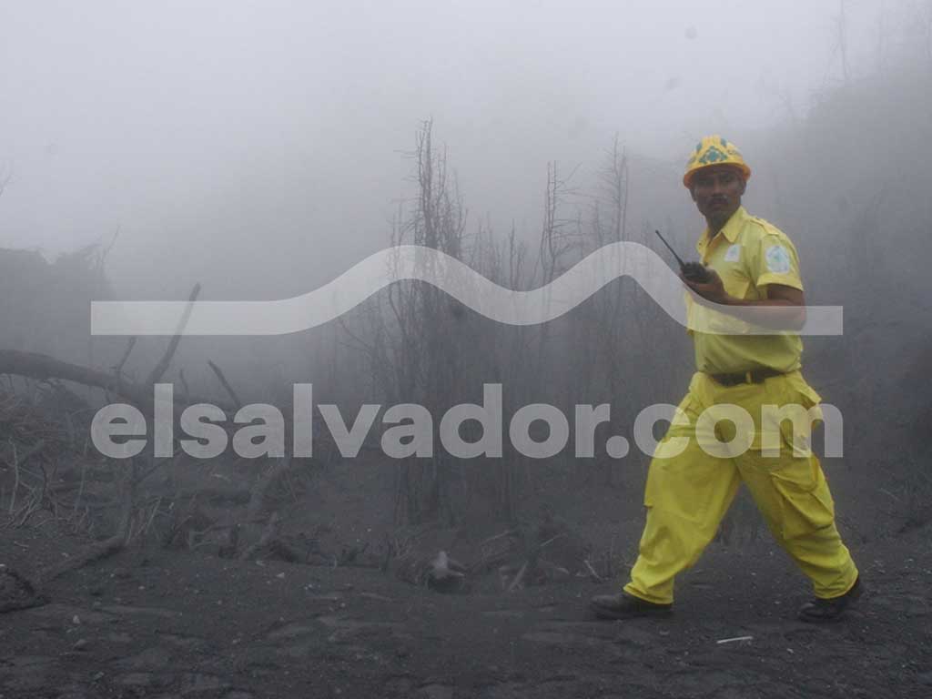 Un hombre con un uniforme amarillo brillante y un casco amarillo camina a través de una niebla gris espesa y ceniza en suspensión, sosteniendo una radio. El fondo está borroso por la niebla.