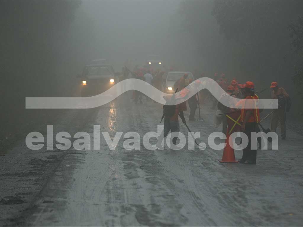 Un grupo de trabajadores de emergencia con uniformes y cascos naranjas caminan por una carretera oscura y cubierta de ceniza húmeda, iluminada por los faros de los vehículos que se aproximan en la distancia.