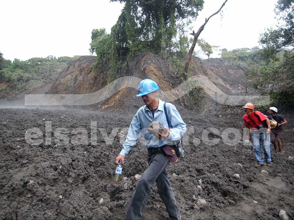 Un hombre con casco azul y camisa azul claro recoge una roca grande de un campo cubierto de lodo gris y ceniza, mientras que otras personas con cascos caminan detrás de él. El fondo muestra una ladera de colina dañada.