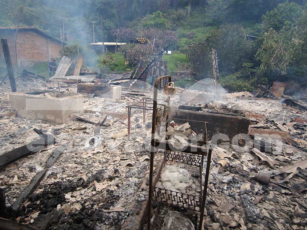 Una vista del interior de una estructura de madera y lámina colapsada y destruida, con escombros carbonizados y ceniza gris esparcidos por el suelo. El fondo muestra vegetación brumosa.