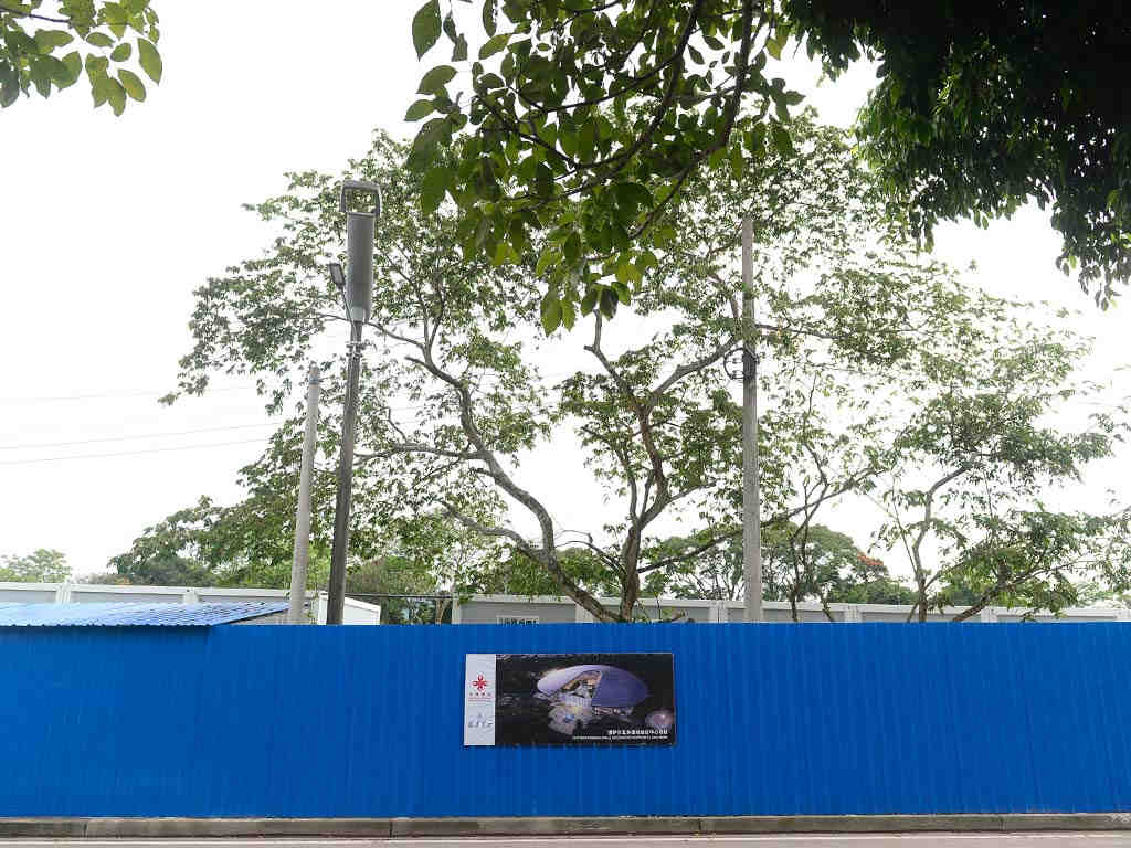 Láminas azules que cercan el predio a lo largo de la calle con un cartel informativo sobre el proyecto frente a árboles altos.