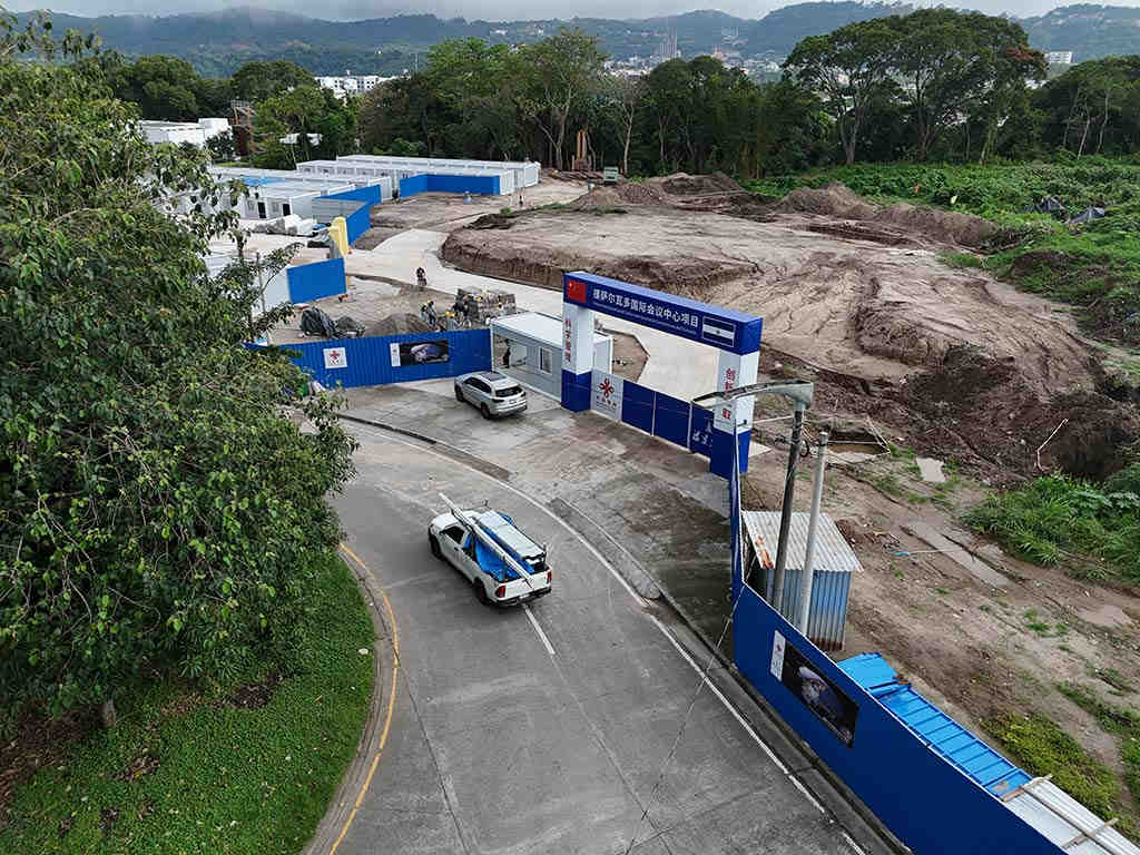 Vista en ángulo del portón de entrada al proyecto con rótulos en chino y español, contenedores de oficinas y la calle de acceso.