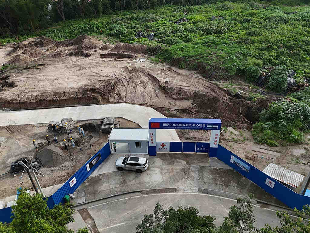 Vista aérea que muestra el portón de entrada azul y blanco del proyecto y las grandes áreas de tierra removida detrás del cerramiento.