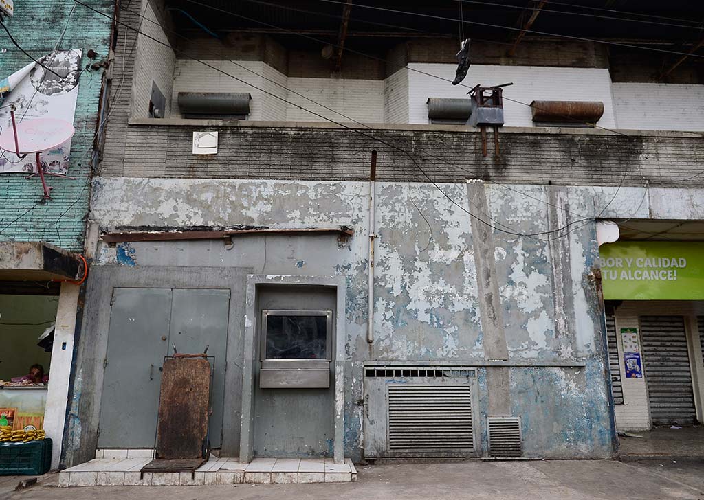 Fotografía a nivel de calle que muestra una pared gris con pintura descascarada, una antigua ventanilla metálica y una puerta tras el retiro de puestos informales.