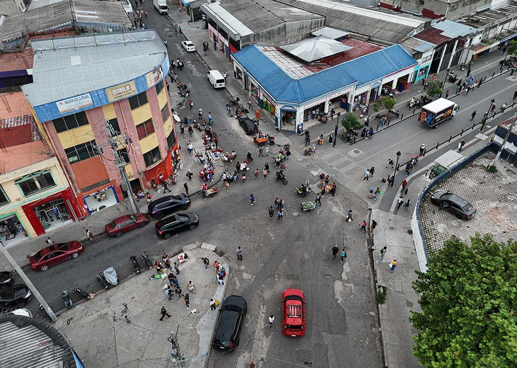 Vista aérea cercana de una esquina con edificios comerciales coloridos, peatones circulando y vehículos transitando por la calle libre.