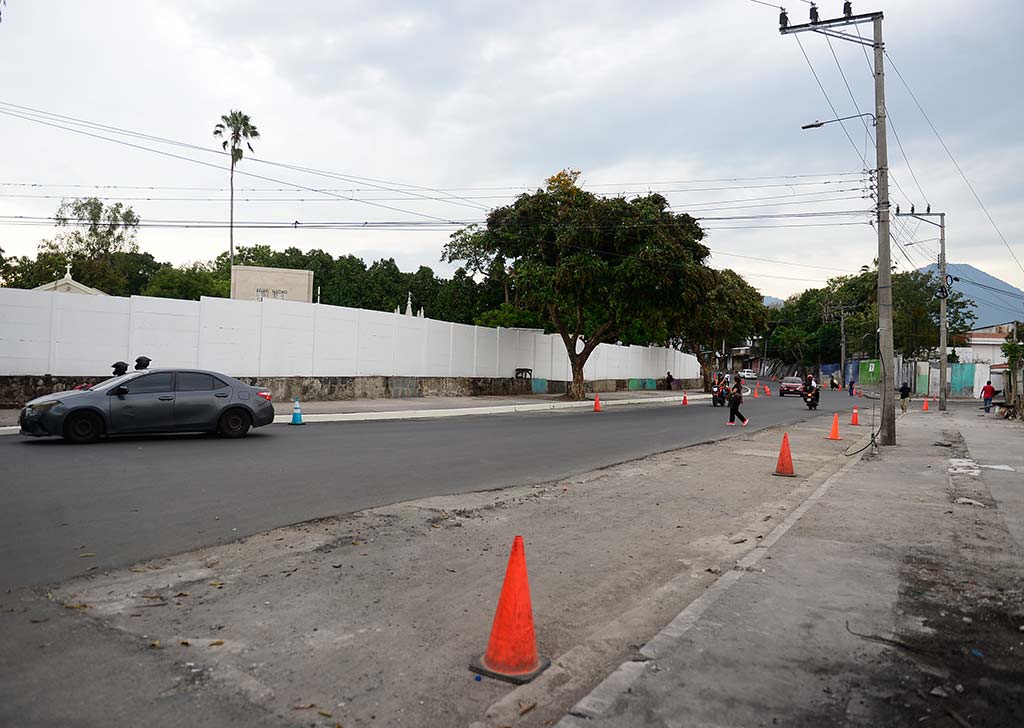 Fotografía a nivel de calle que muestra una carretera de asfalto despejada con conos de señalización color naranja a la derecha y el muro blanco del Cementerio Los Ilustres a la izquierda.
