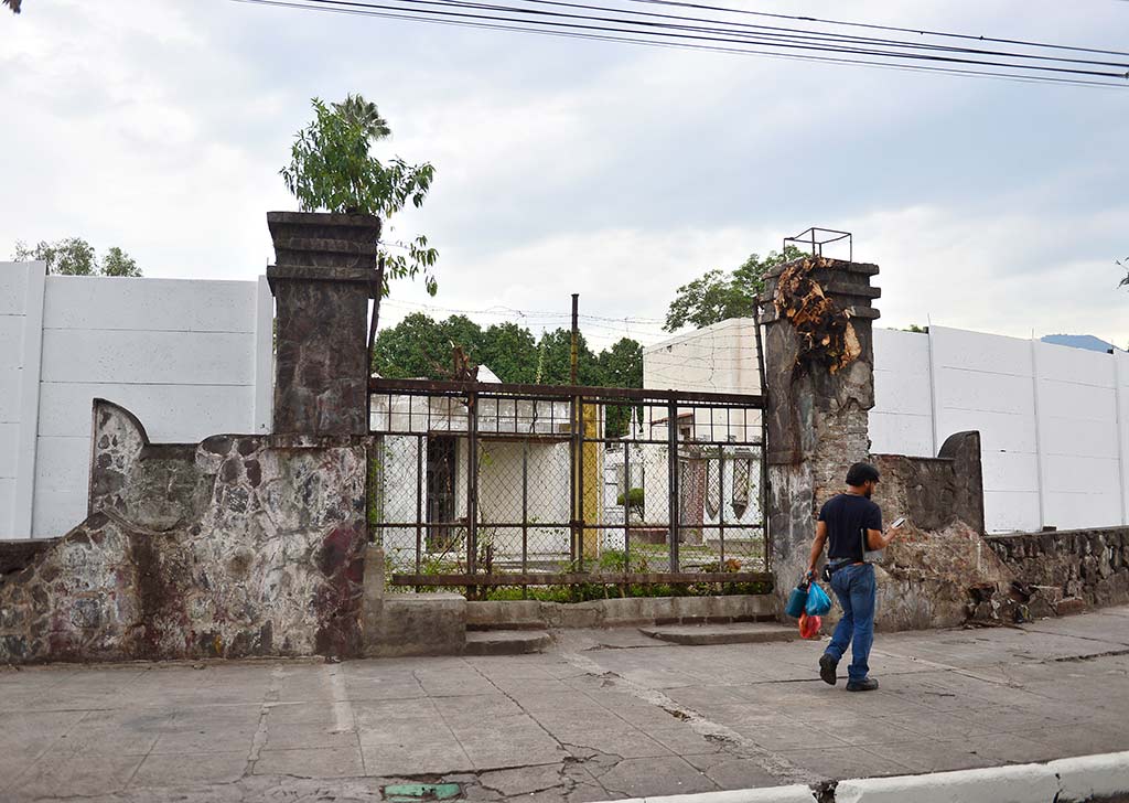 Un peatón caminando frente a una antigua estructura de portón de piedra y hierro enmarcada por muros blancos en una acera limpia.