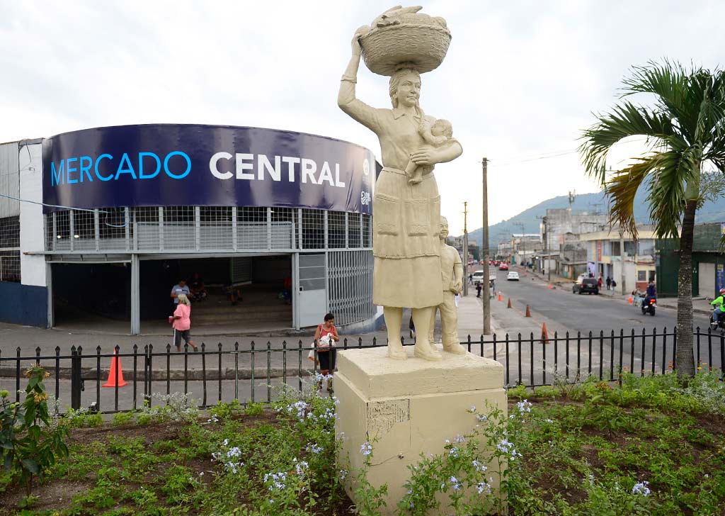 Estatua de color crema de una mujer con una canasta en la cabeza y un niño en brazos, ubicada en un arriate ajardinado frente al edificio del Mercado Central.