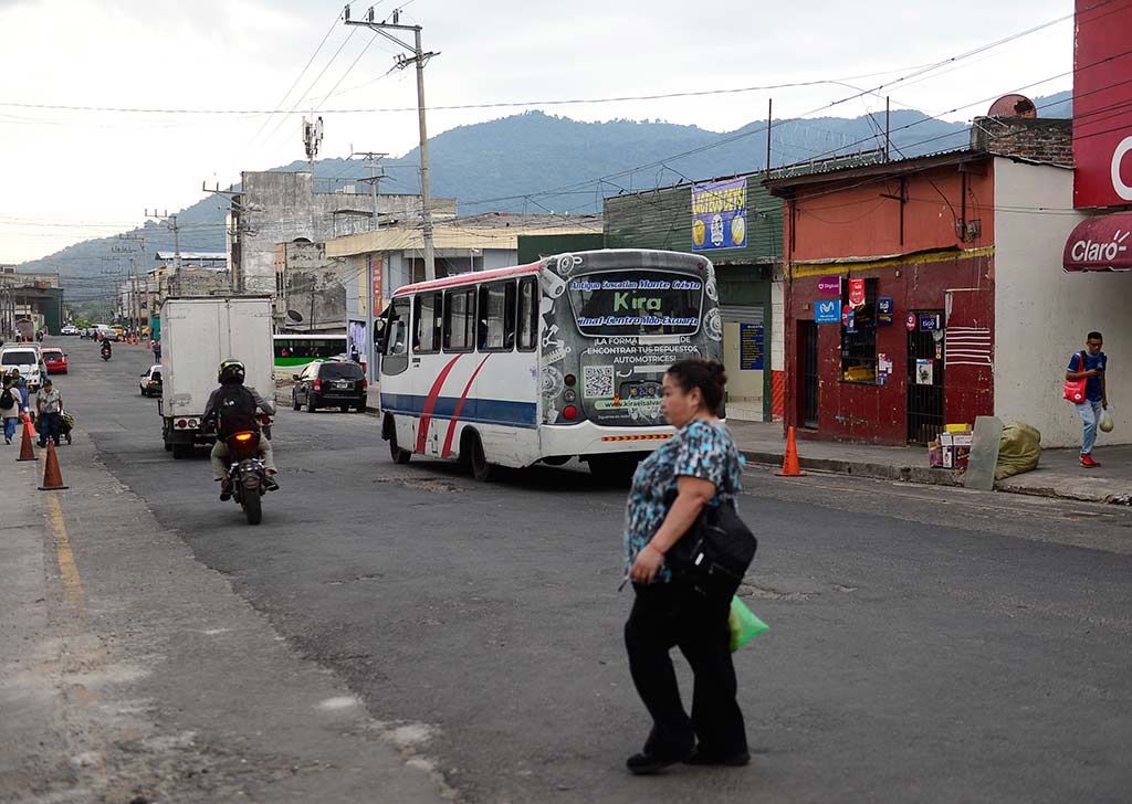 Una mujer caminando en primer plano mientras un microbús del transporte público transita por una calle despejada con conos de señalización.