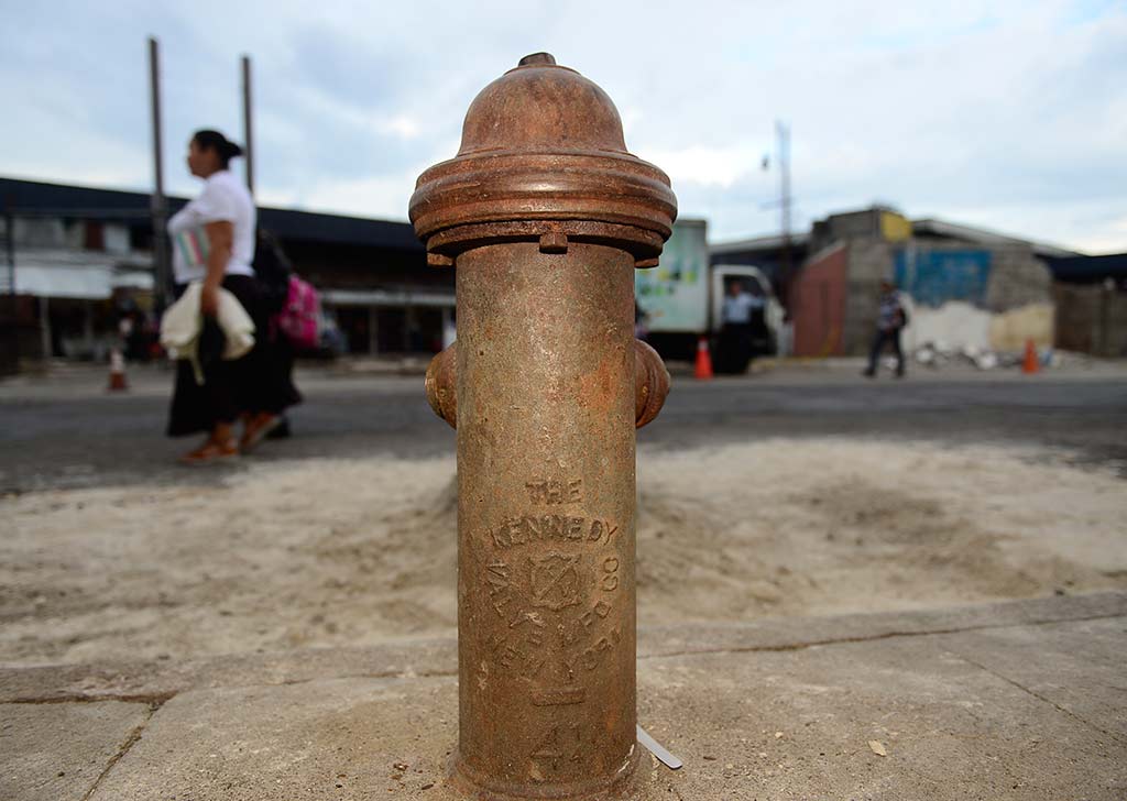 Primer plano de un hidrante metálico antiguo y oxidado sobre una acera despejada con transeúntes desenfocados al fondo