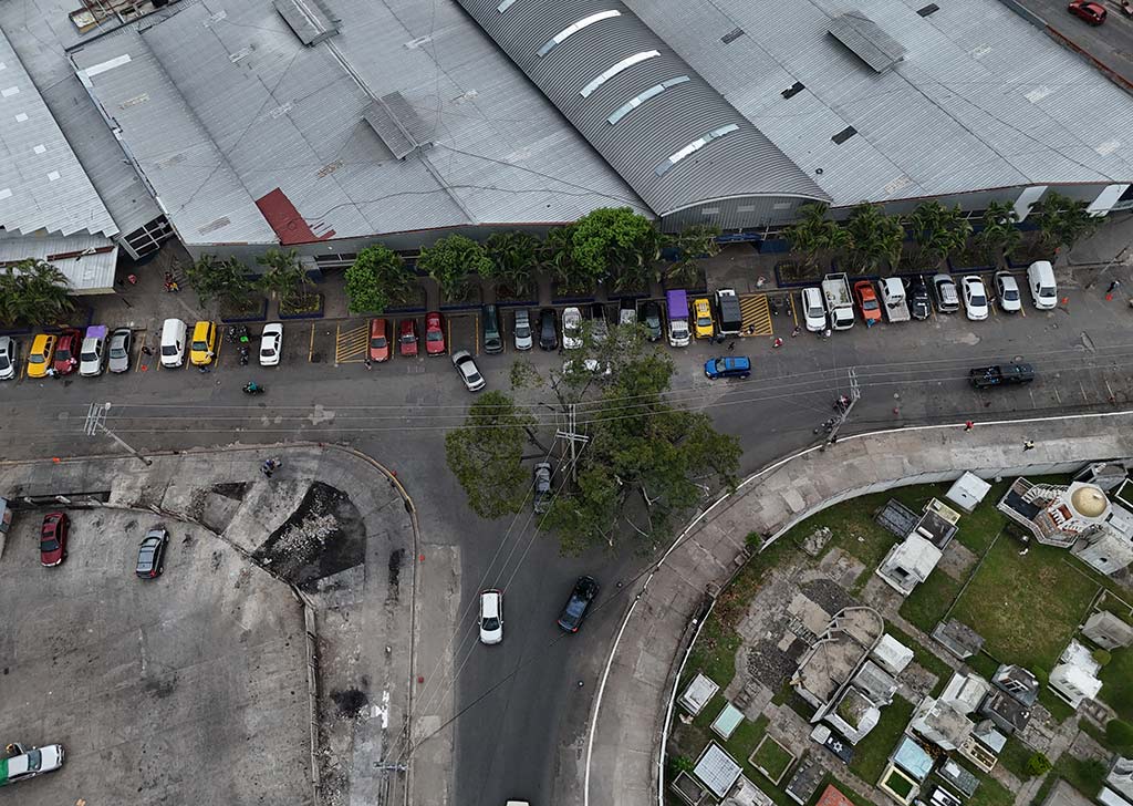 Vista aérea que muestra una calle despejada con vehículos estacionados en fila entre un edificio con techo arqueado y las tumbas de un cementerio.