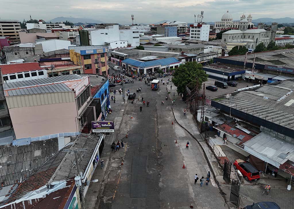 Vista aérea a media altura de una avenida despejada con algunos peatones y farmacias, con la cúpula de Catedral al fondo.