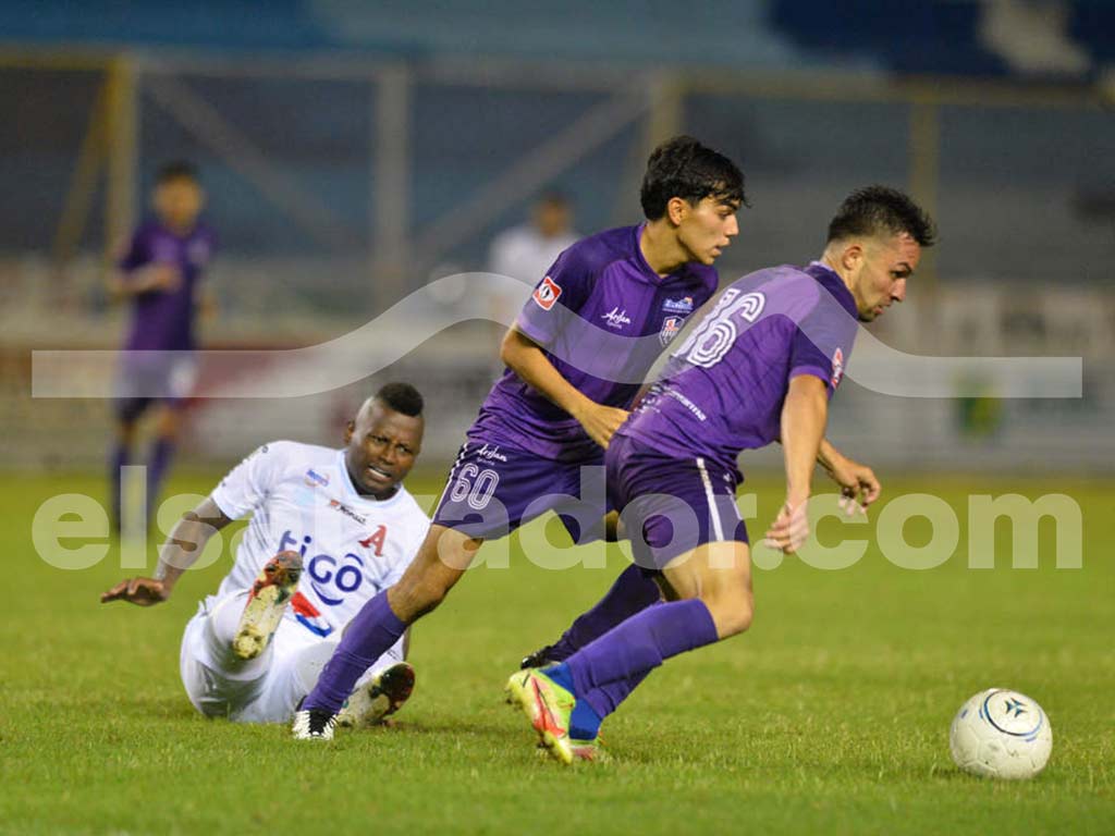Una foto a color de un jugador de AD Chalatenango con su uniforme azul y blanco, corriendo con el balón en un estadio soleado con montañas de fondo.