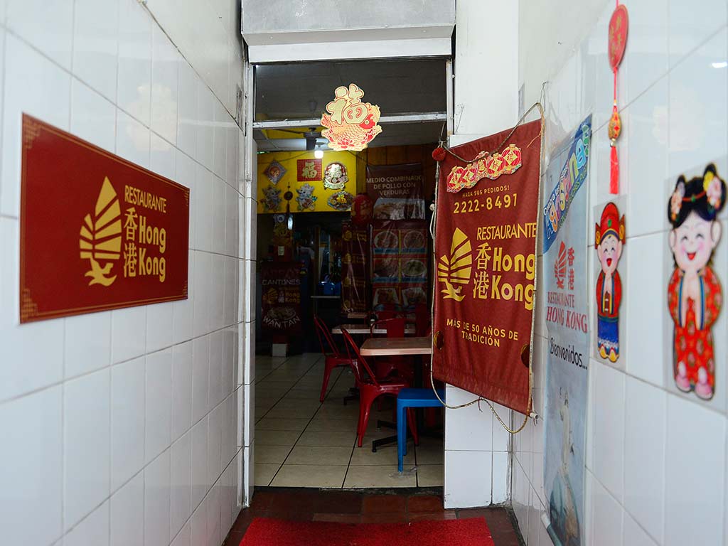Vista de un pasillo estrecho con paredes de baldosas blancas que lleva al interior de un restaurante. El pasillo está decorado con varios elementos chinos, incluyendo un letrero rojo de "Restaurante Hong Kong", faroles rojos colgantes, carteles con caracteres chinos dorados y un banderín rojo. Al fondo, se ve el comedor con mesas y sillas rojas, y más decoraciones en las paredes amarillas.
