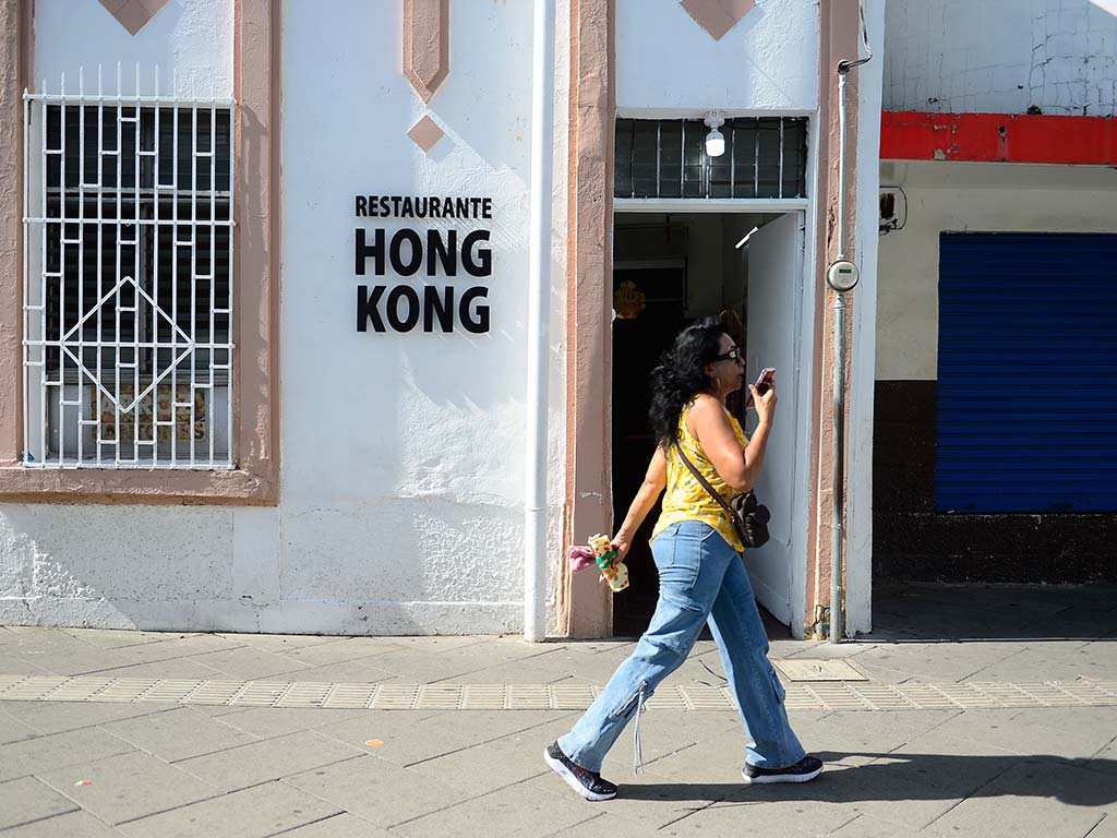 Una mujer con una camiseta amarilla y vaqueros azules camina rápidamente por la acera soleada. Detrás de ella, la fachada blanca y rosa de un edificio histórico con un gran rótulo negro que dice "RESTAURANTE HONG KONG". La puerta está abierta, revelando un interior oscuro.