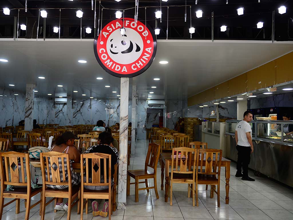Vista interior de un restaurante chino bien iluminado con techos altos y baldosas blancas. Mesas y sillas de madera están dispuestas ordenadamente. Al fondo, comensales sentados disfrutan de su comida. Un gran cartel circular colgante con un oso panda y texto que dice "ASIA FOOD COMIDA CHINA" cuelga del techo. Un empleado con una camiseta blanca está de pie cerca de la barra de servicio.