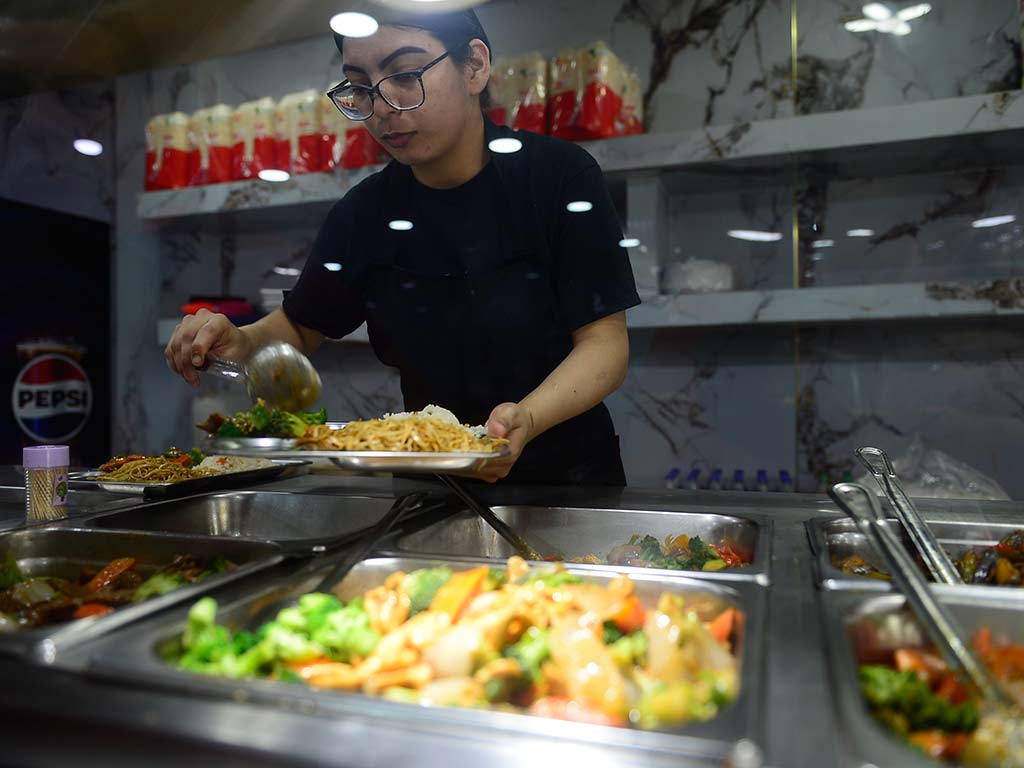 Una joven empleada con gafas y una camiseta negra está de pie detrás de una barra de servicio de acero inoxidable con varios recipientes de comida caliente. Ella está sirviendo una porción de chao mein humeante en un plato de metal con una cuchara grande. El fondo está desenfocado, mostrando más bandejas de comida.