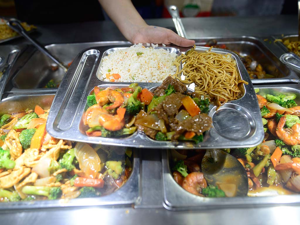 Primer plano de una bandeja de metal con cuatro compartimentos llena de comida china. Un compartimento tiene arroz frito, otro chao mein, un tercero pollo agridulce con verduras y el cuarto brócoli salteado. La bandeja está sostenida por una mano y el fondo muestra más bandejas de comida en la barra de servicio.