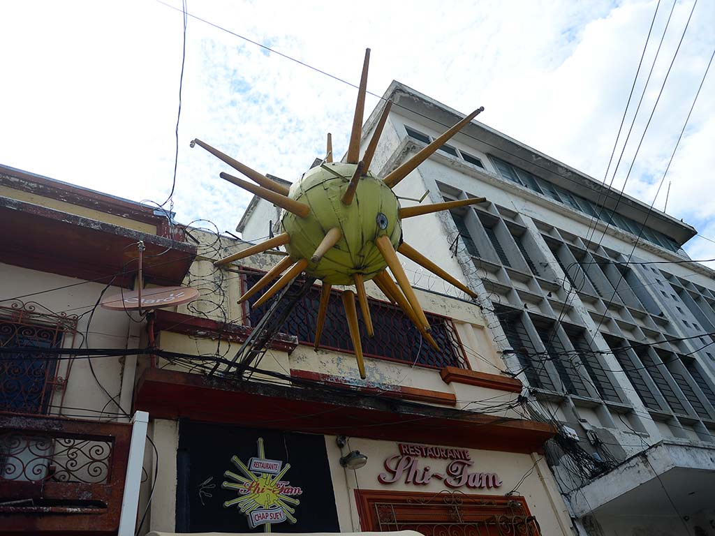 Vista en ángulo hacia arriba de la fachada de un edificio antiguo con cableado eléctrico desordenado. Un gran cartel de estrella amarilla con puntas de madera sobresale sobre la entrada del "RESTAURANTE SHI-TAM". Debajo, hay un letrero más pequeño con el nombre del restaurante y "CHAP SUEY". El edificio es de color blanco y marrón con ventanas enrejadas. El cielo está nublado.