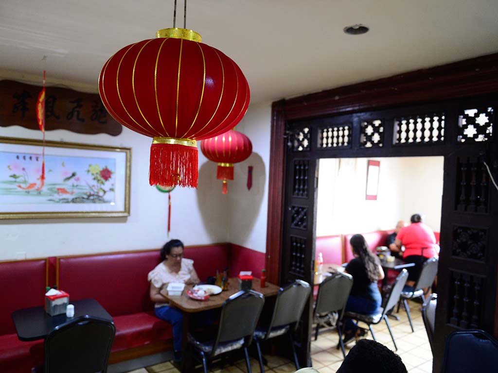 Vista interior de un restaurante chino con iluminación cálida. Un gran farol rojo de seda cuelga del techo en primer plano. Mesas y sillas de metal están ocupadas por personas comiendo. Las paredes blancas tienen decoraciones chinas y cabinas rojas. El suelo es de baldosas.