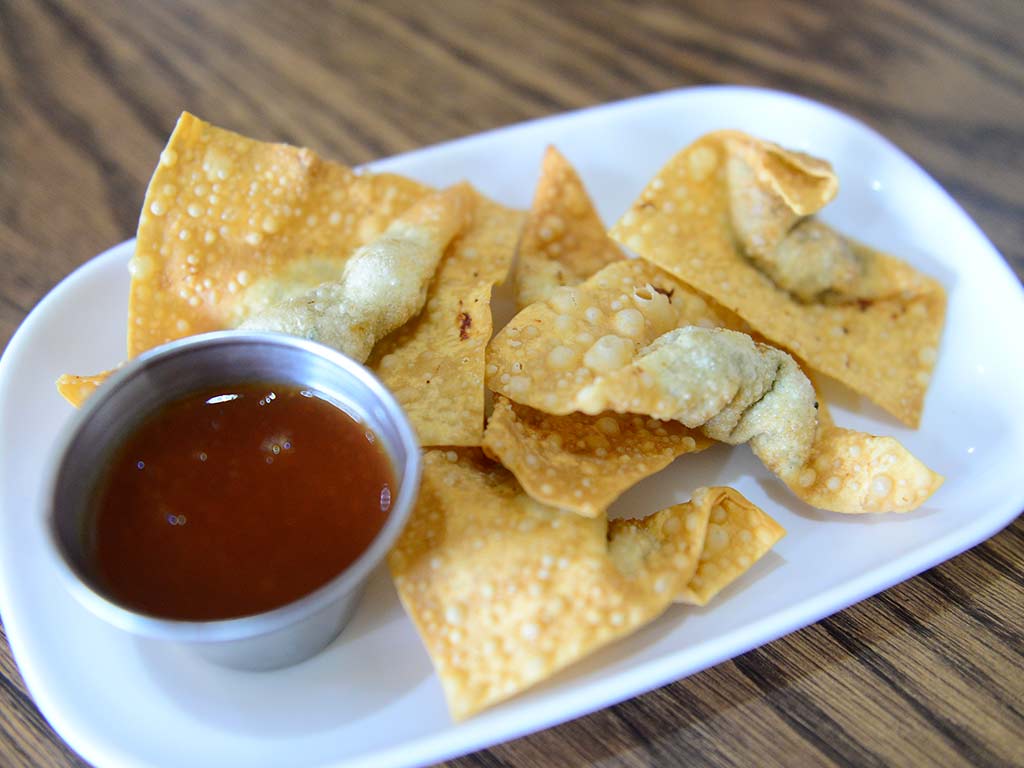 Primer plano de un plato blanco con varios wantanes fritos, dorados y crujientes. Al lado, un pequeño tazón de metal con salsa agridulce de color naranja. El plato está sobre una mesa de madera con un fondo desenfocado.