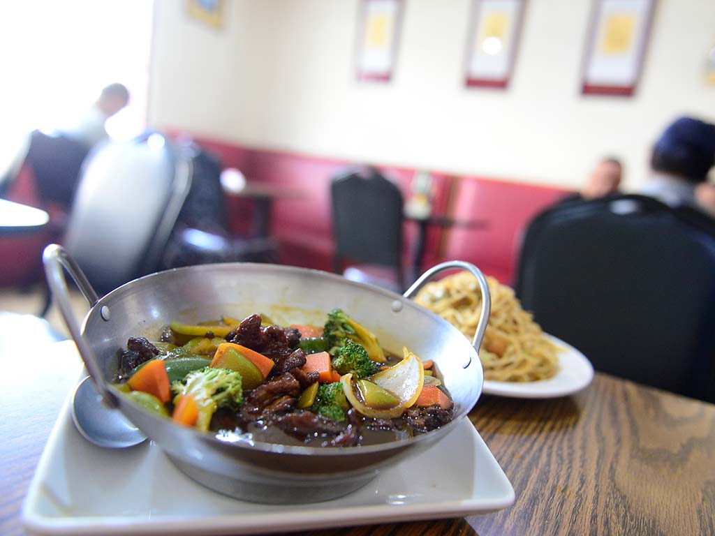 Primer plano de un wok metálico lleno de carne salteada, brócoli, zanahorias y pimientos en una salsa oscura y espesa. De fondo desenfocado, un plato blanco con chao mein y un panecillo. La escena está sobre una mesa de madera en un restaurante.