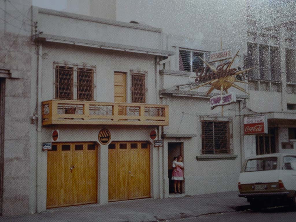 Fotografía de estilo antiguo de una calle urbana. Muestra un edificio de dos pisos pintado de blanco con un balcón de madera marrón y dos puertas de garaje de madera en la planta baja. Un letrero luminoso de "RESTAURANTE" con letras chinas y un rótulo de "Coca-Cola" cuelgan en la esquina. Un coche antiguo está aparcado a la derecha. Una persona está de pie en una puerta estrecha.