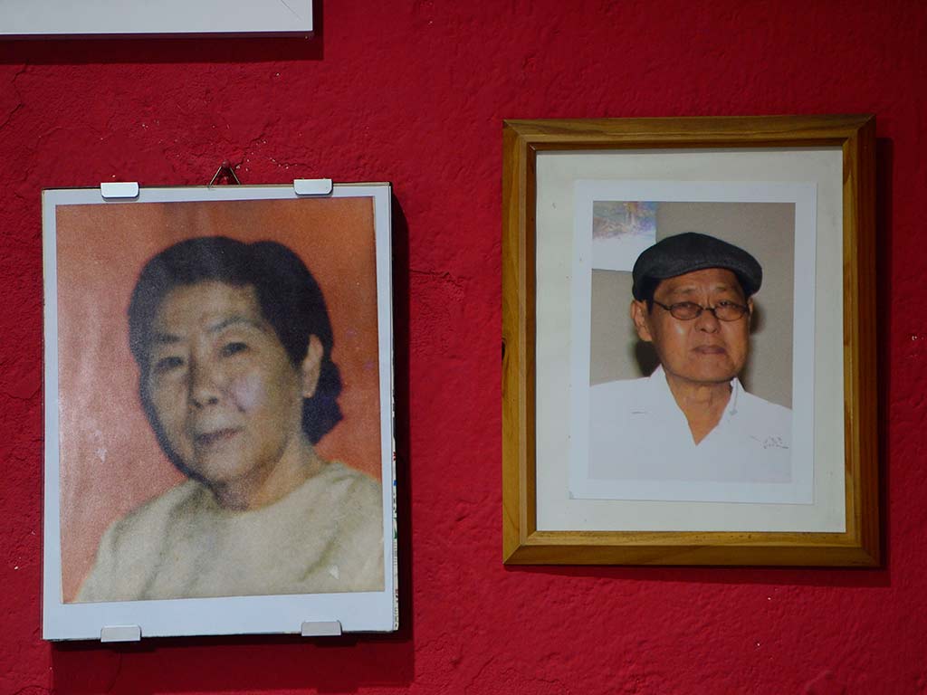 Dos fotografías enmarcadas y colgadas sobre una pared de color rojo intenso. A la izquierda, un retrato antiguo de una mujer asiática con cabello oscuro y expresión serena. A la derecha, un retrato de un hombre asiático mayor con gafas y una gorra negra, ambos con ropa clara.