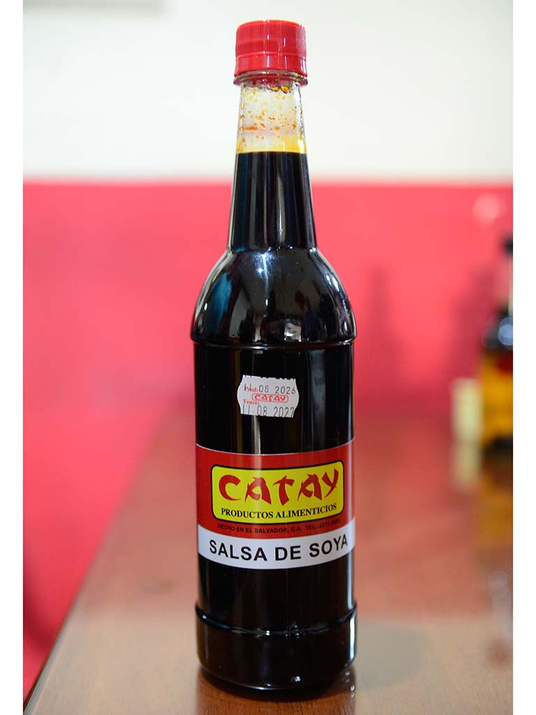 Primer plano de una botella de vidrio transparente llena de salsa de soya oscura, de pie sobre una mesa de madera en un restaurante. La botella tiene una etiqueta roja y amarilla que dice "CATAY Productos Alimenticios HECHO EN EL SALVADOR SALSA DE SOYA". El fondo es un panel de pared rojo desenfocado.